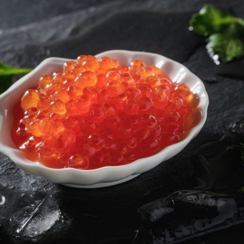 Red caviar in the white bowl in seashell shape on black wet shale board with ice.